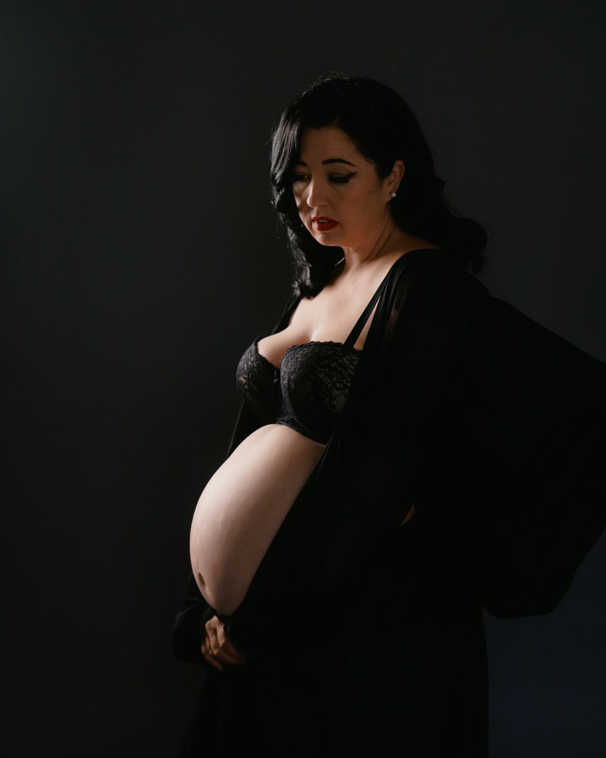Pregnant woman in black lingerie and sheer robe cradling baby bump against dark studio background.