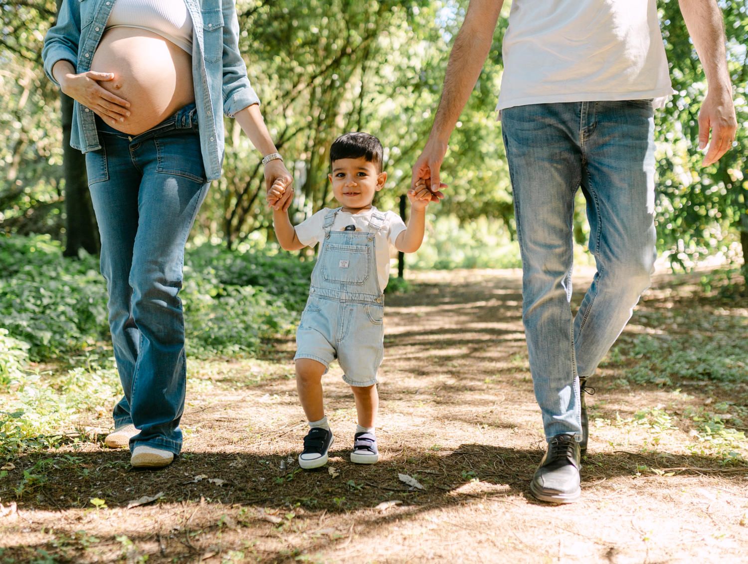 Pregnant woman and partner walking with toddler on a forest path, holding hands in a family maternity photo.