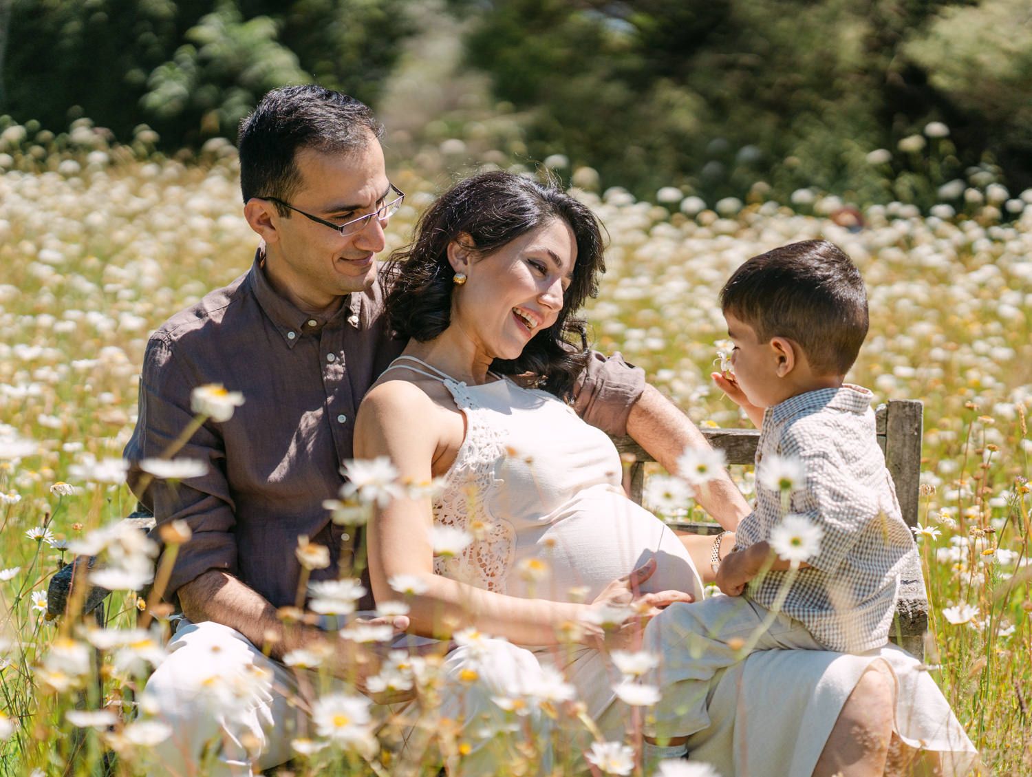 Pregnant woman sits with husband and young son in a sunny daisy field for a family maternity photo session.