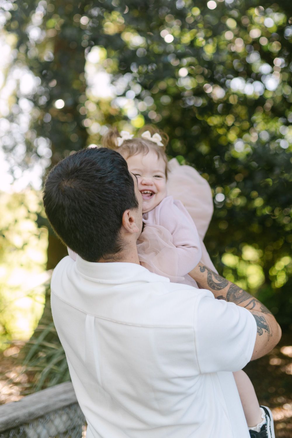 A tattooed father holds his laughing baby girl in a pink outfit outdoors surrounded by lush green trees.