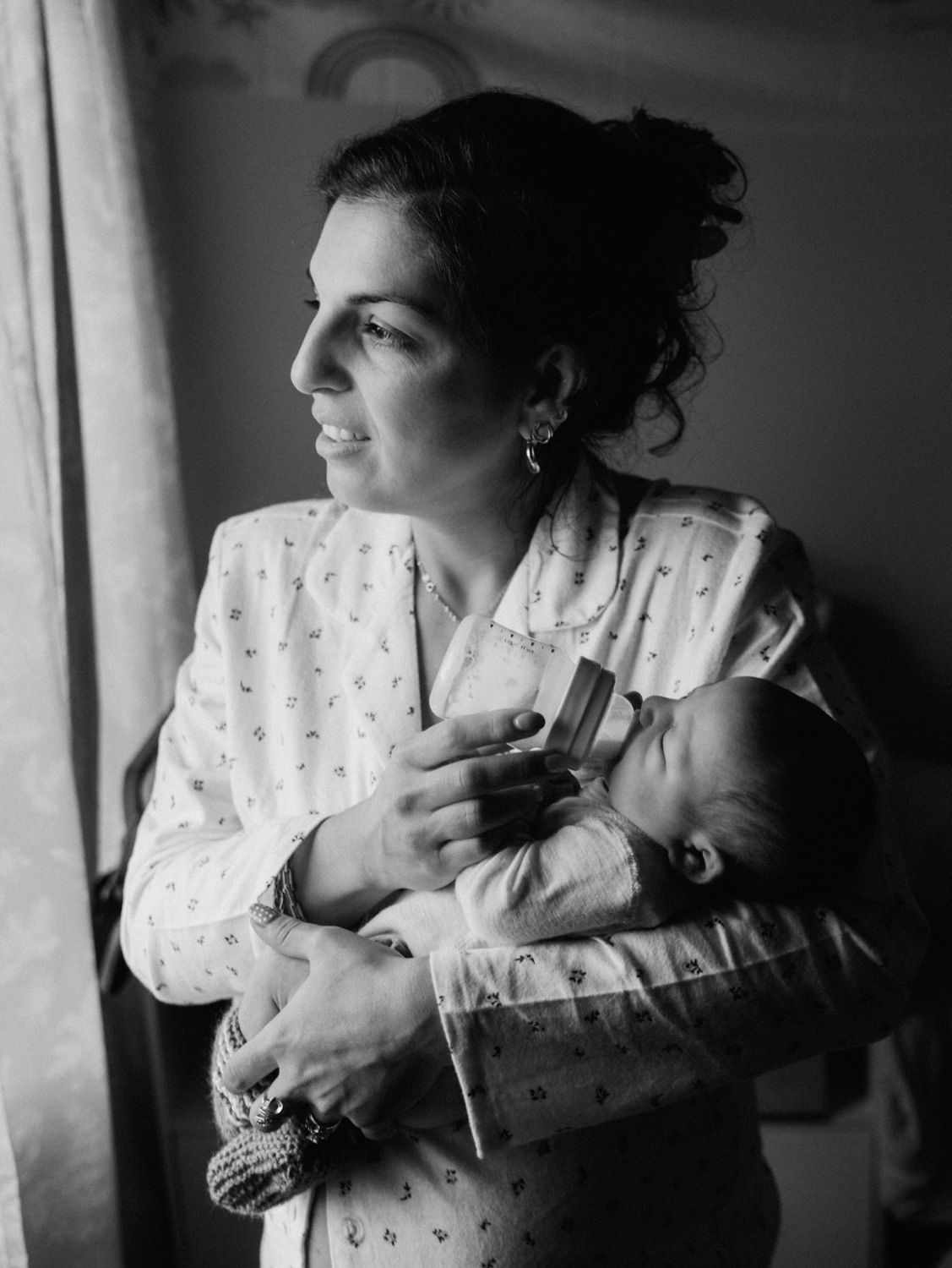 Black and white photo of a mother in a floral gown tenderly holding her newborn baby near a bright window.