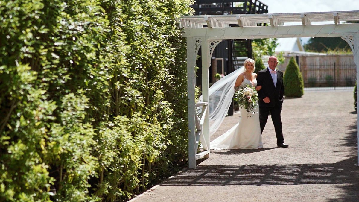Bride in white gown and veil walks with escort through garden pergola on sunny wedding day.