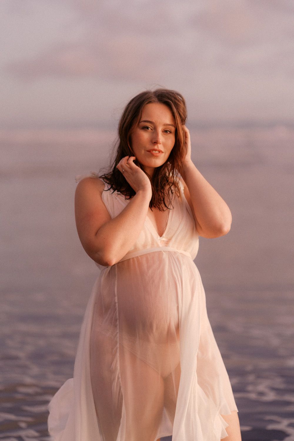 Pregnant woman in sheer white dress posing on beach at golden sunset, hands in hair.