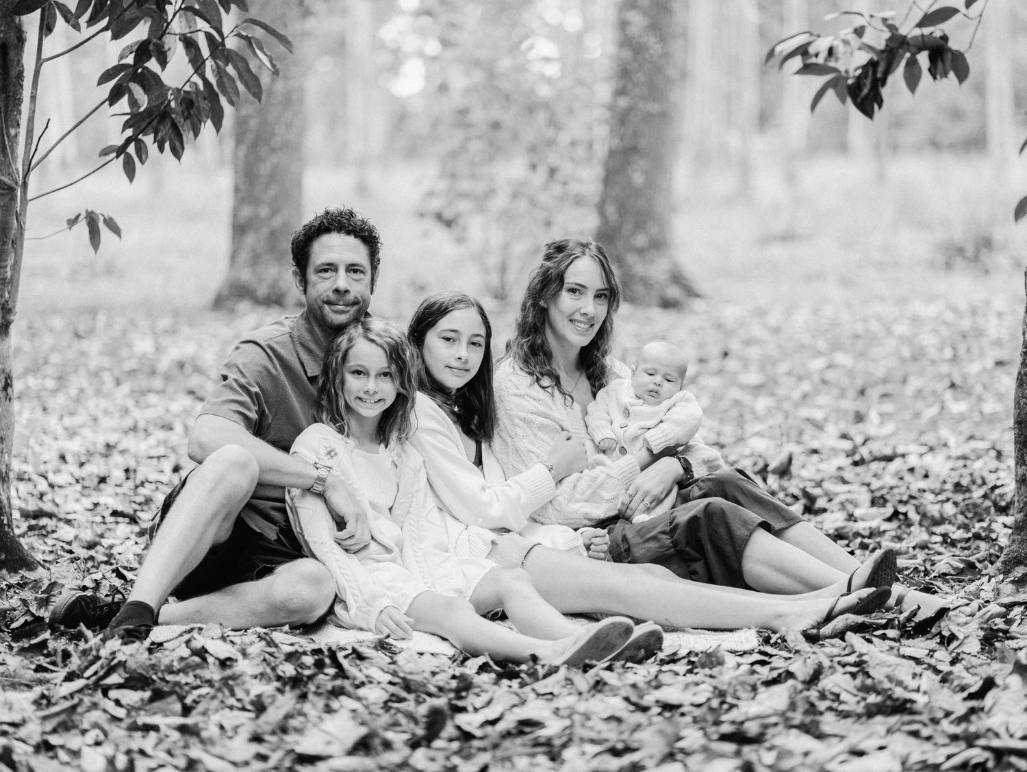 Black and white family portrait of parents with three children sitting on fallen leaves in a forest setting.