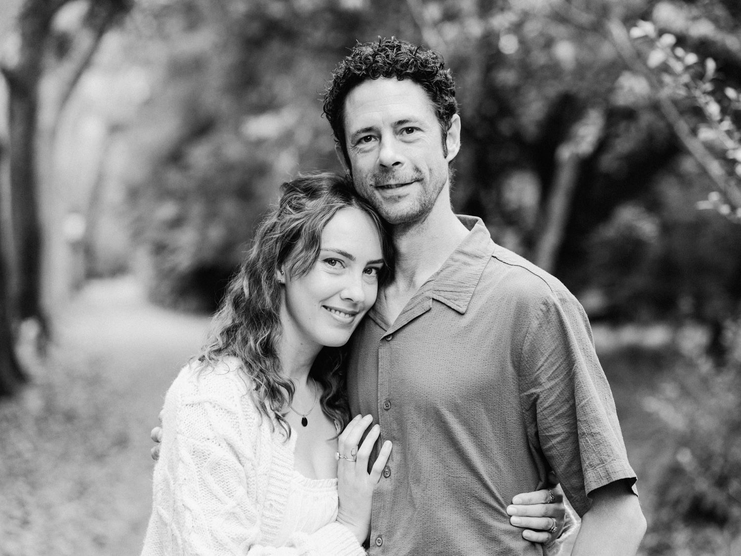 Black and white portrait of a smiling couple embracing outdoors among lush trees in a park setting.