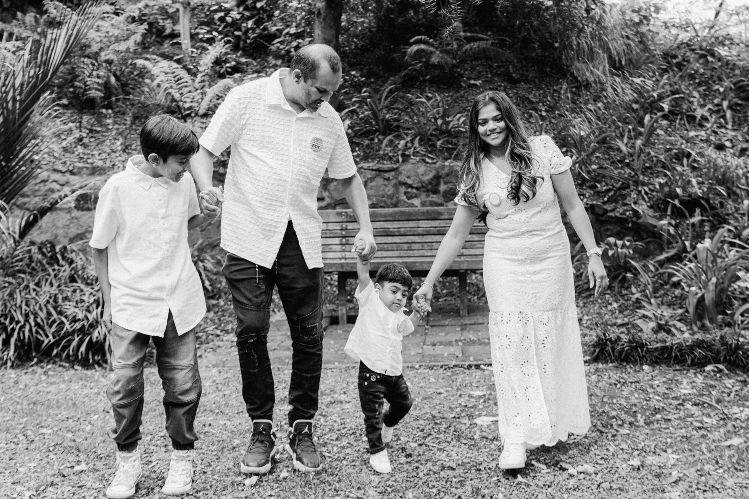 Black and white family photo of parents swinging toddler while older child walks beside them in lush garden.