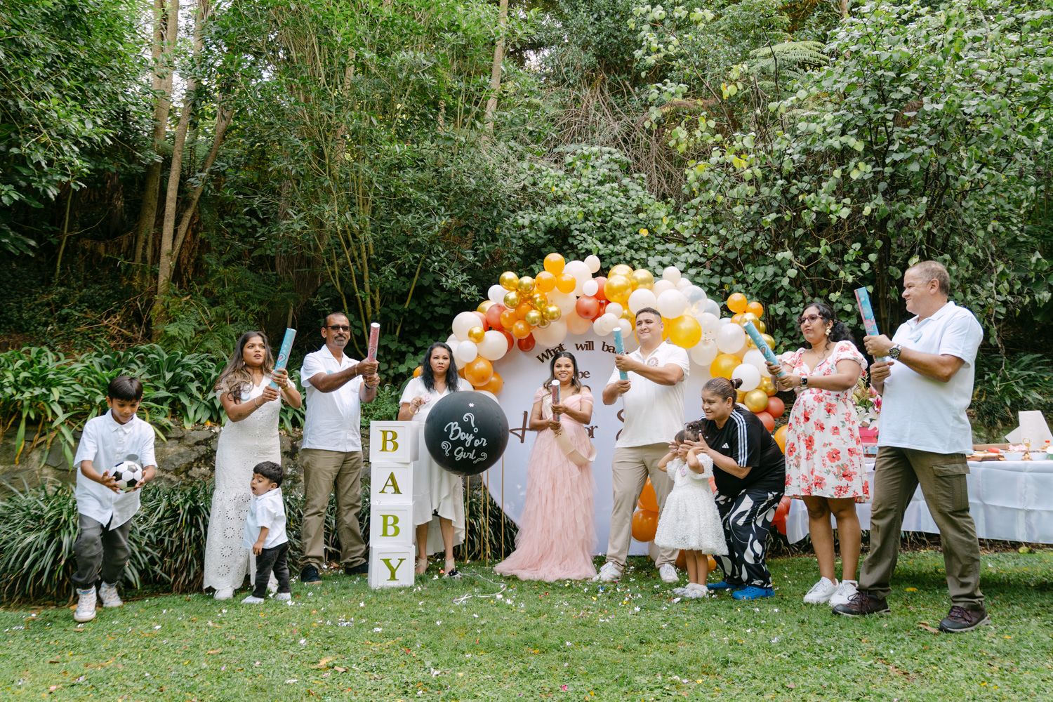 Group of family and friends celebrating a baby shower outdoors with gold balloons arch and BABY sign in lush garden.