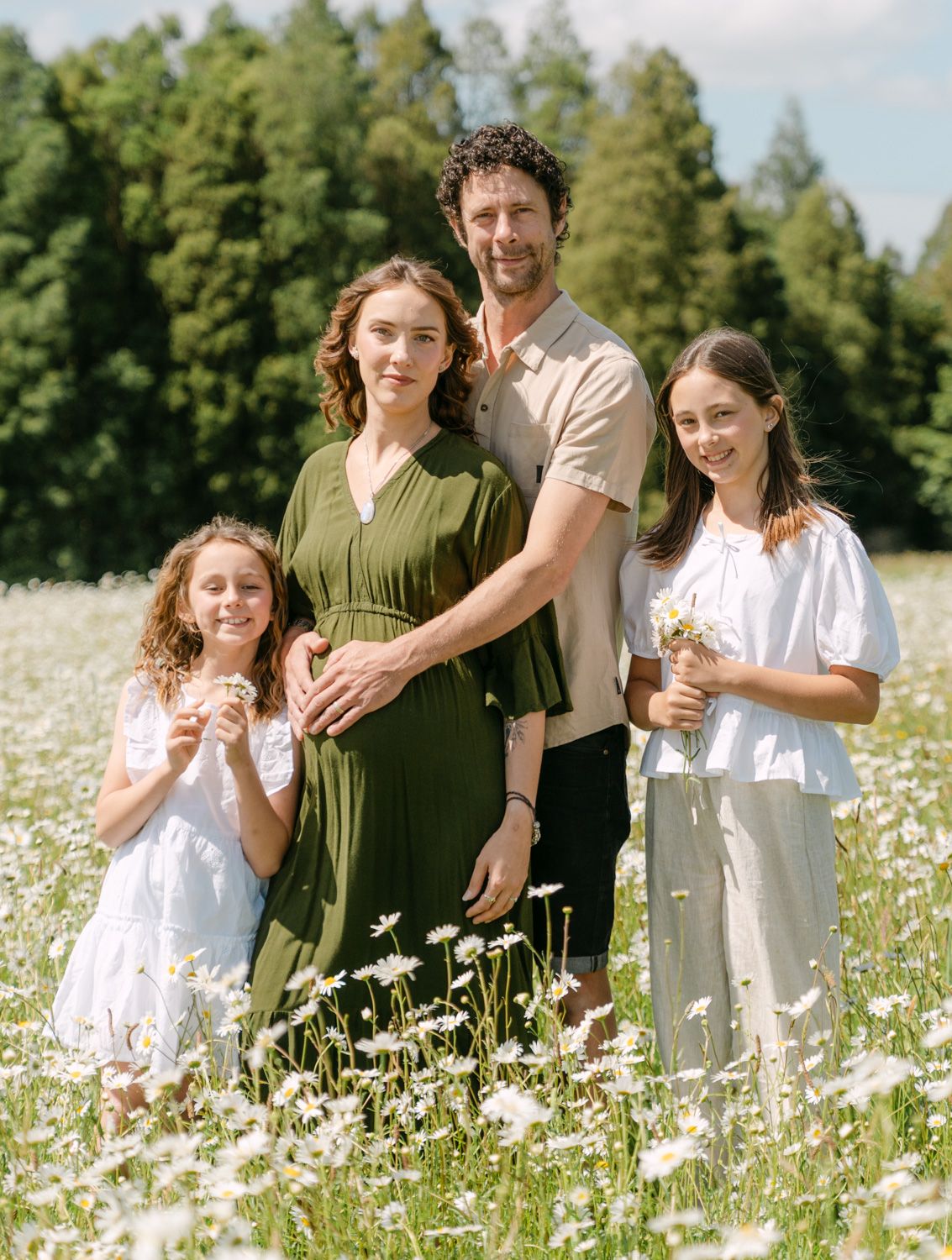Family of four posing in a sunny daisy field, two girls in white dresses with parents, surrounded by green trees.