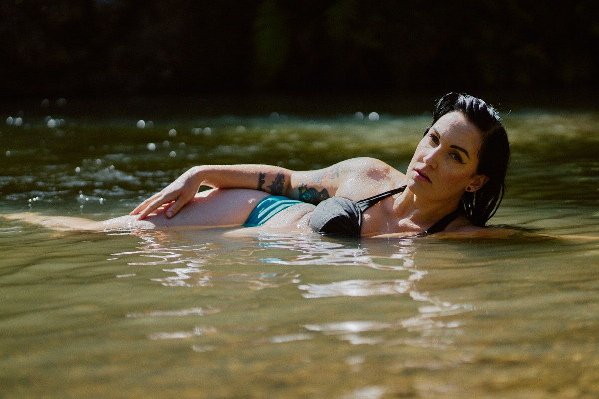 Woman in teal bikini floating in a sunlit natural river, showcasing tattoos and wet hair.