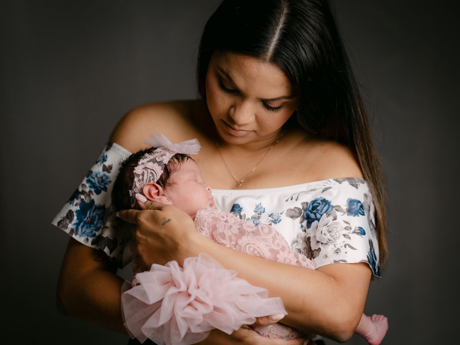 A mother in a floral off-shoulder dress tenderly holds her newborn baby girl dressed in a pink tutu headband.