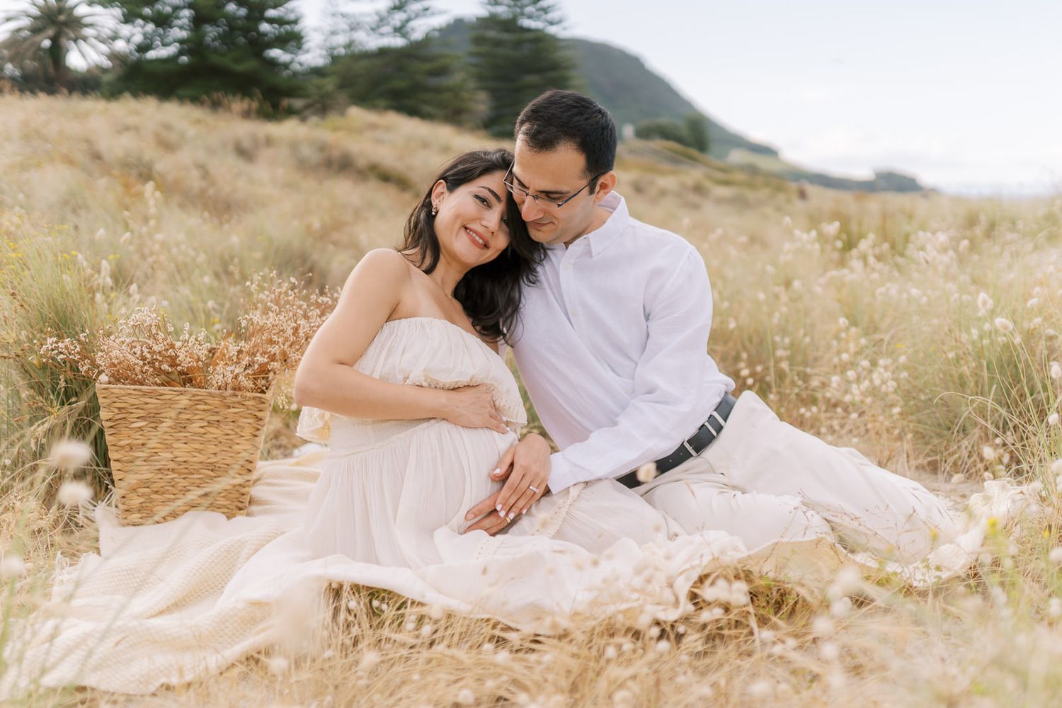 Couple sitting in golden field, man tenderly embracing pregnant woman in white dress during maternity photoshoot.