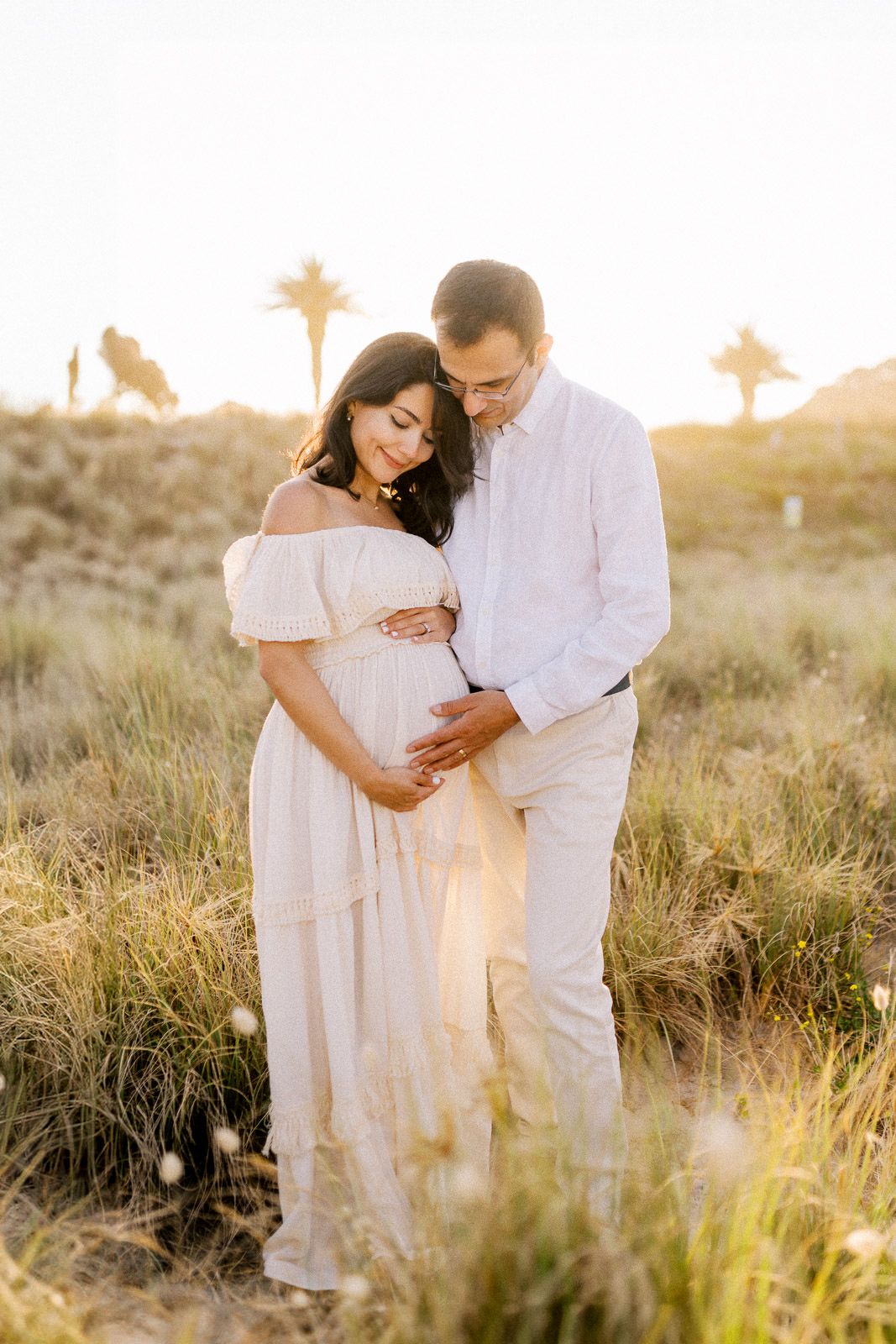 Couple in white outfits embracing outdoors at golden hour, pregnant woman cradling baby bump in sunlit grassy field.