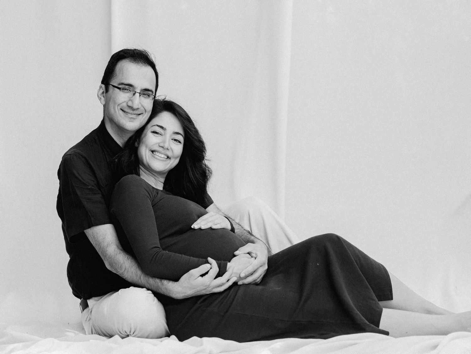 Black and white maternity photo of smiling couple sitting together, man embracing pregnant woman in dark dress.