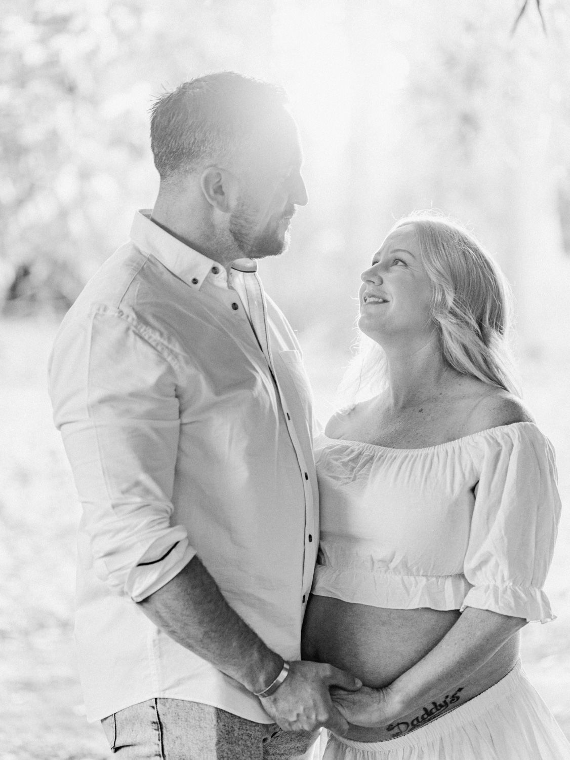 Black and white maternity photo of a couple holding hands outdoors, woman in off-shoulder dress smiling up at bearded man.