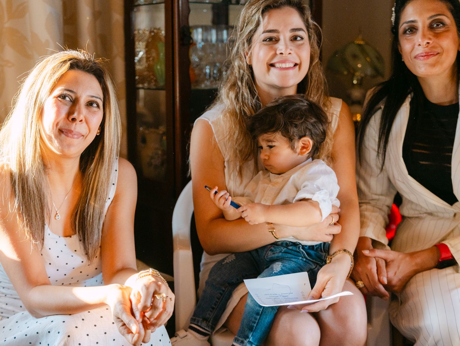 Three women sitting together, middle woman holding a baby, smiling warmly in a cozy indoor setting.