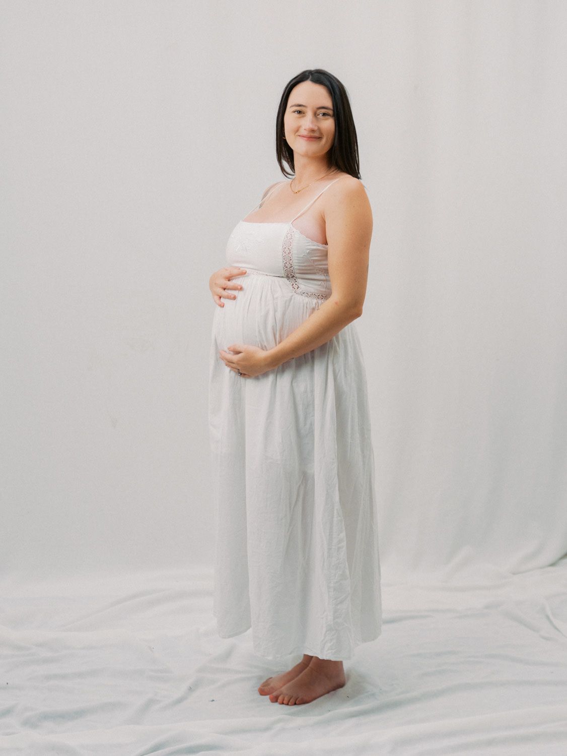 Pregnant woman in white maxi dress cradling baby bump, posing barefoot against a white studio background.