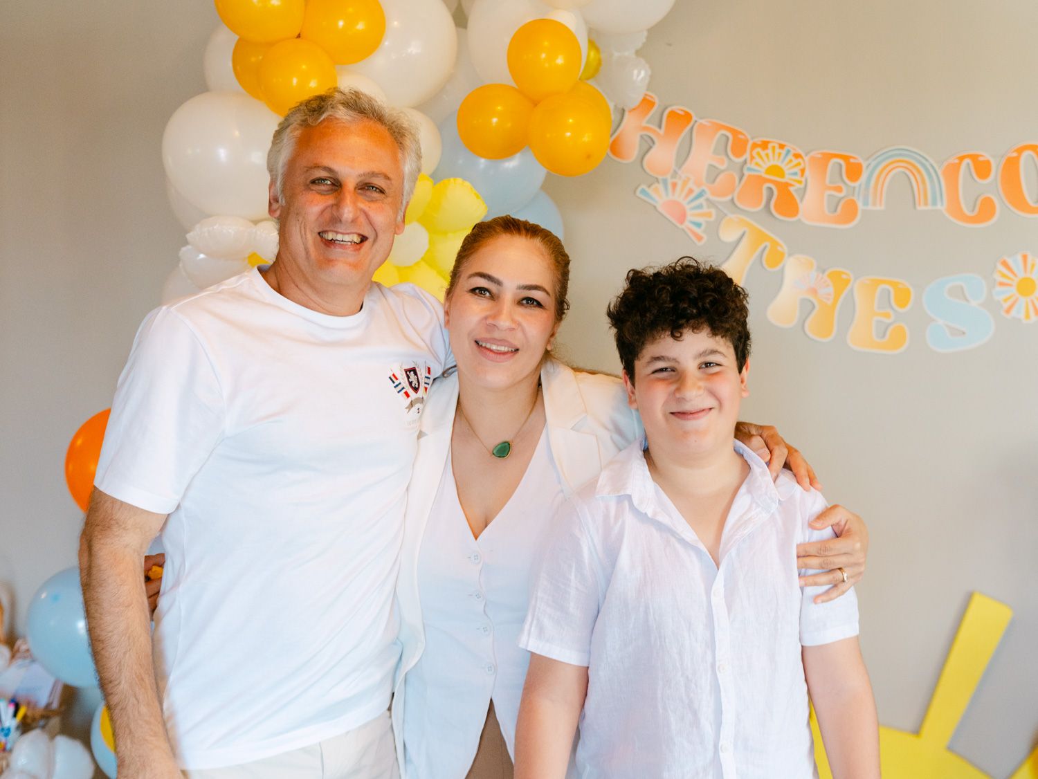 Three people smiling at a festive party with yellow and white balloon decorations in the background.