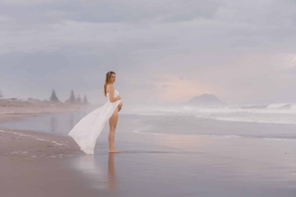 Woman in flowing white dress standing on a serene beach at sunset with gentle waves and misty skies.