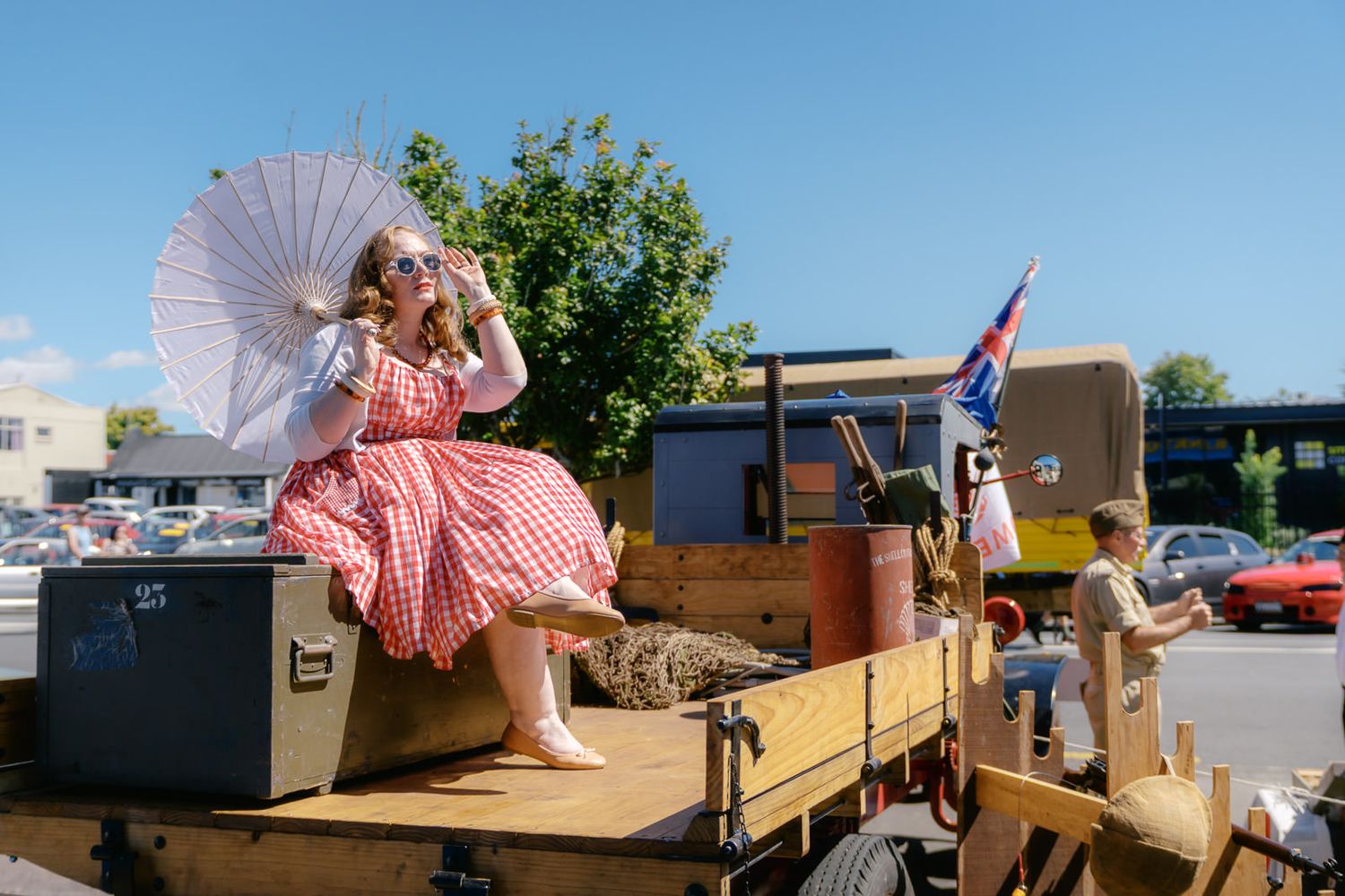 Woman in pink dress holding white parasol sits on a wooden parade float under a sunny blue sky.