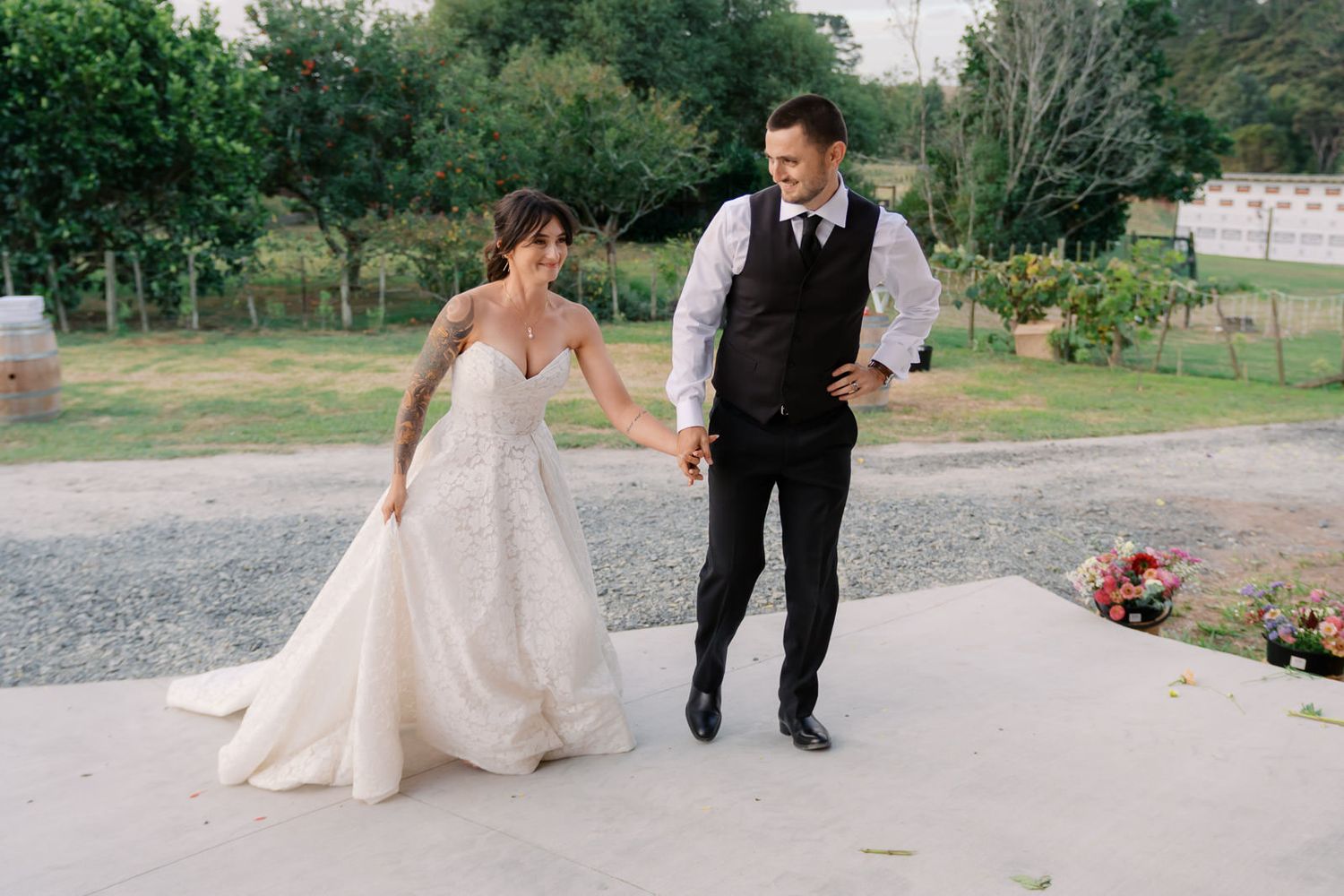 Bride in lace gown and groom in black suit holding hands, walking together outdoors at a garden wedding venue.