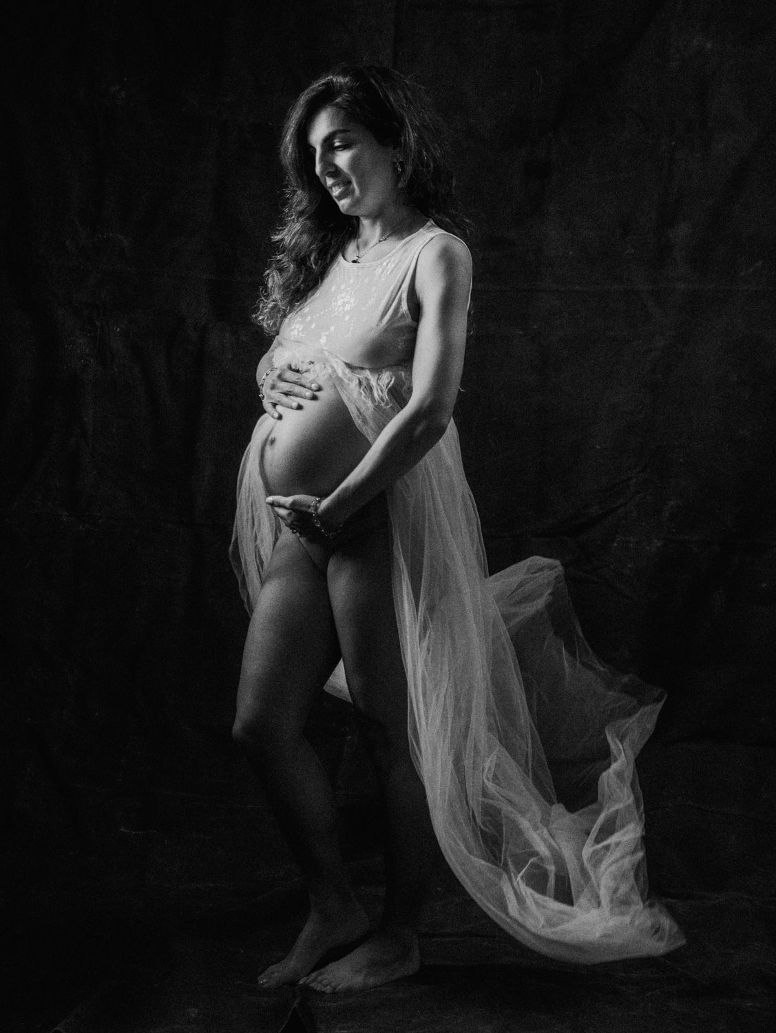 Black and white maternity portrait of a pregnant woman in flowing sheer gown, cradling her baby bump in a studio setting.