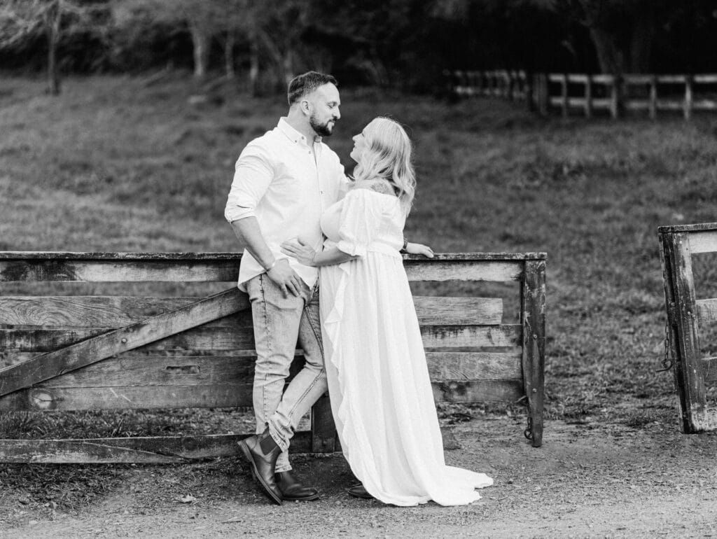 Black and white maternity photo of couple leaning against wooden fence in countryside.