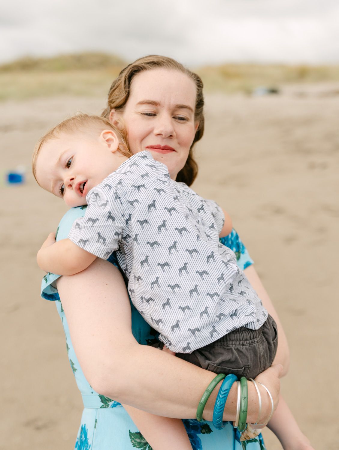 A mother tenderly holds her toddler on a sandy beach, both dressed casually, with dunes in the soft background.