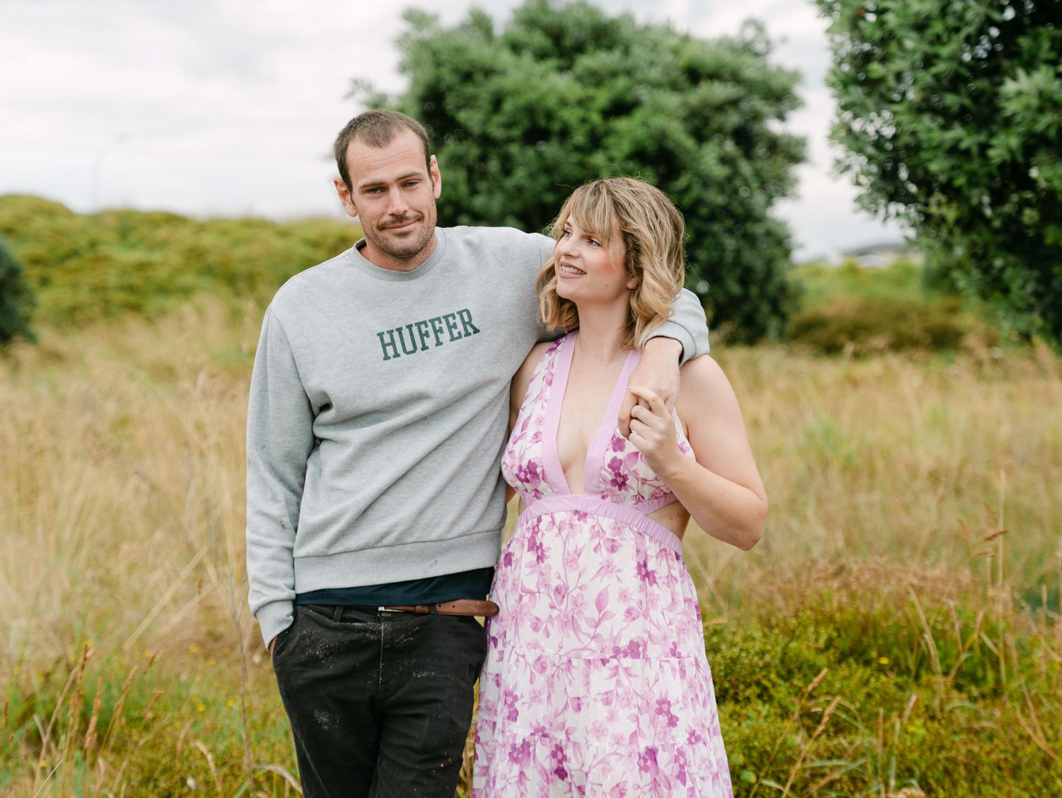Couple smiling together in a field, man in Huffer sweatshirt, woman in floral pink dress.