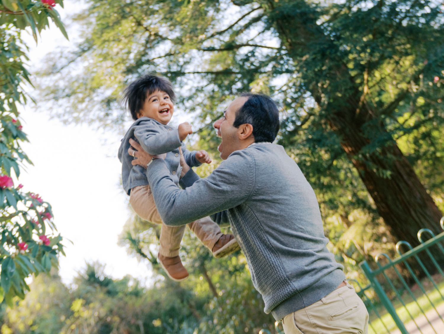 A joyful father lifts his laughing toddler in a sunny park surrounded by lush green trees and red flowers.