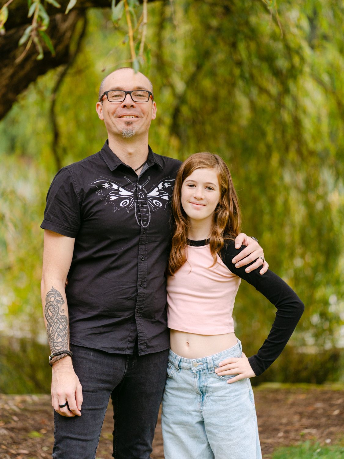 A bald man with tattoos in a black shirt poses with a young girl in a pink crop top and jeans outdoors.