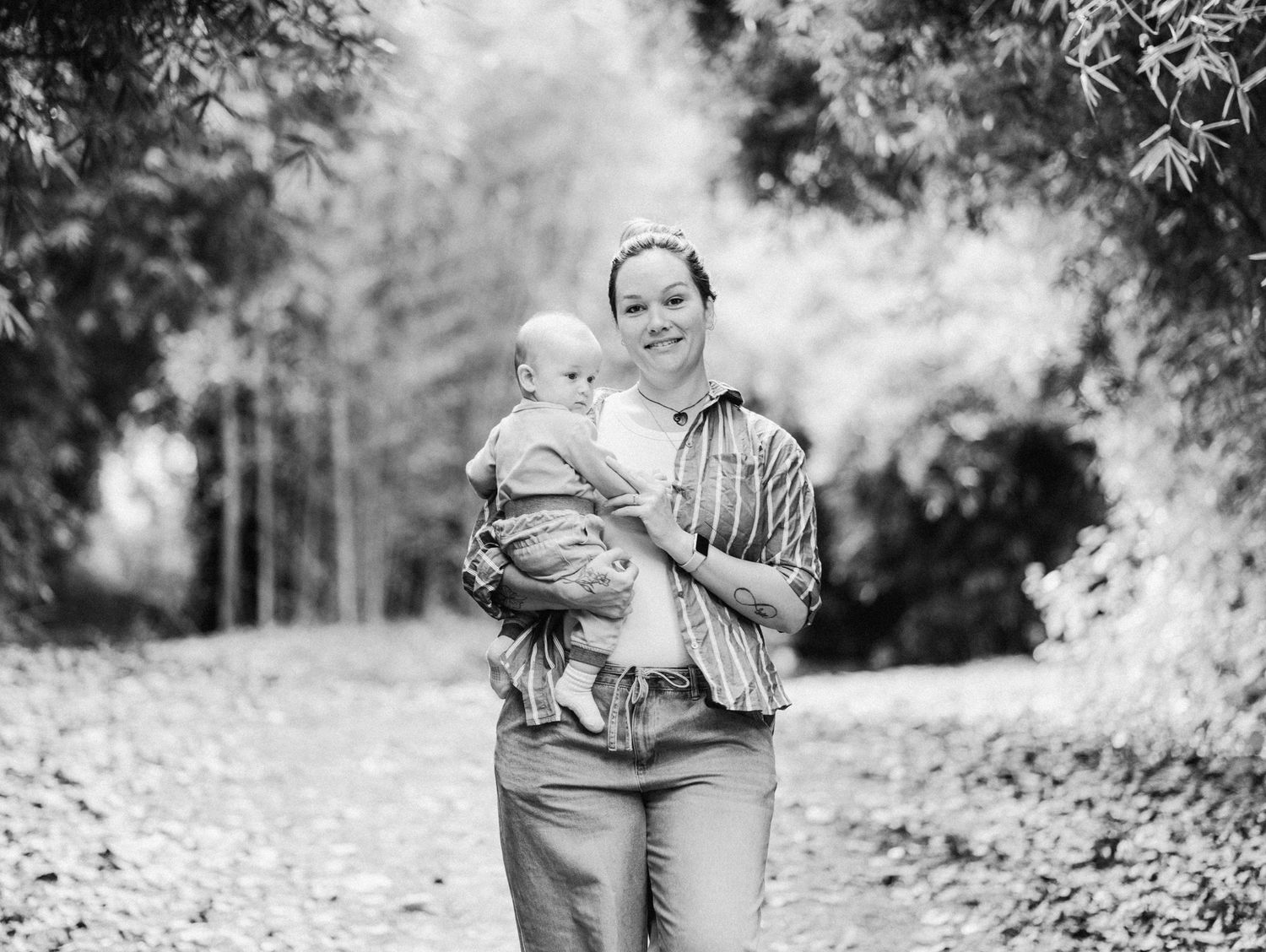 Black and white photo of a smiling woman holding a baby on a tree-lined path outdoors.