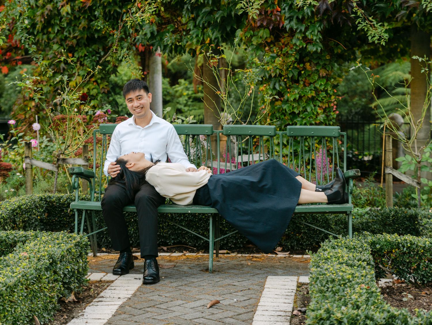 Couple relaxing on a green garden bench surrounded by lush hedges and colorful foliage in a scenic park.