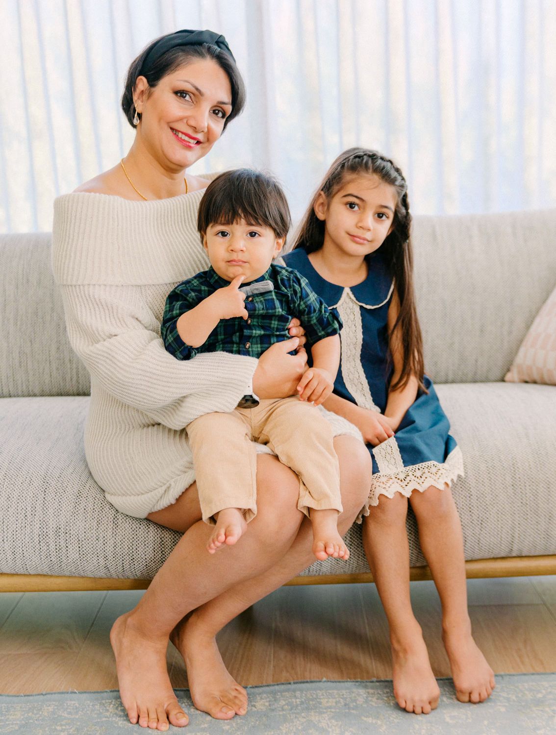 A woman in a cream sweater dress sits on a sofa smiling, holding two young children dressed in navy and plaid.