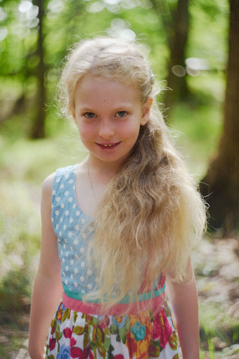 Young blonde girl in colorful floral dress smiling in a sunlit forest setting.