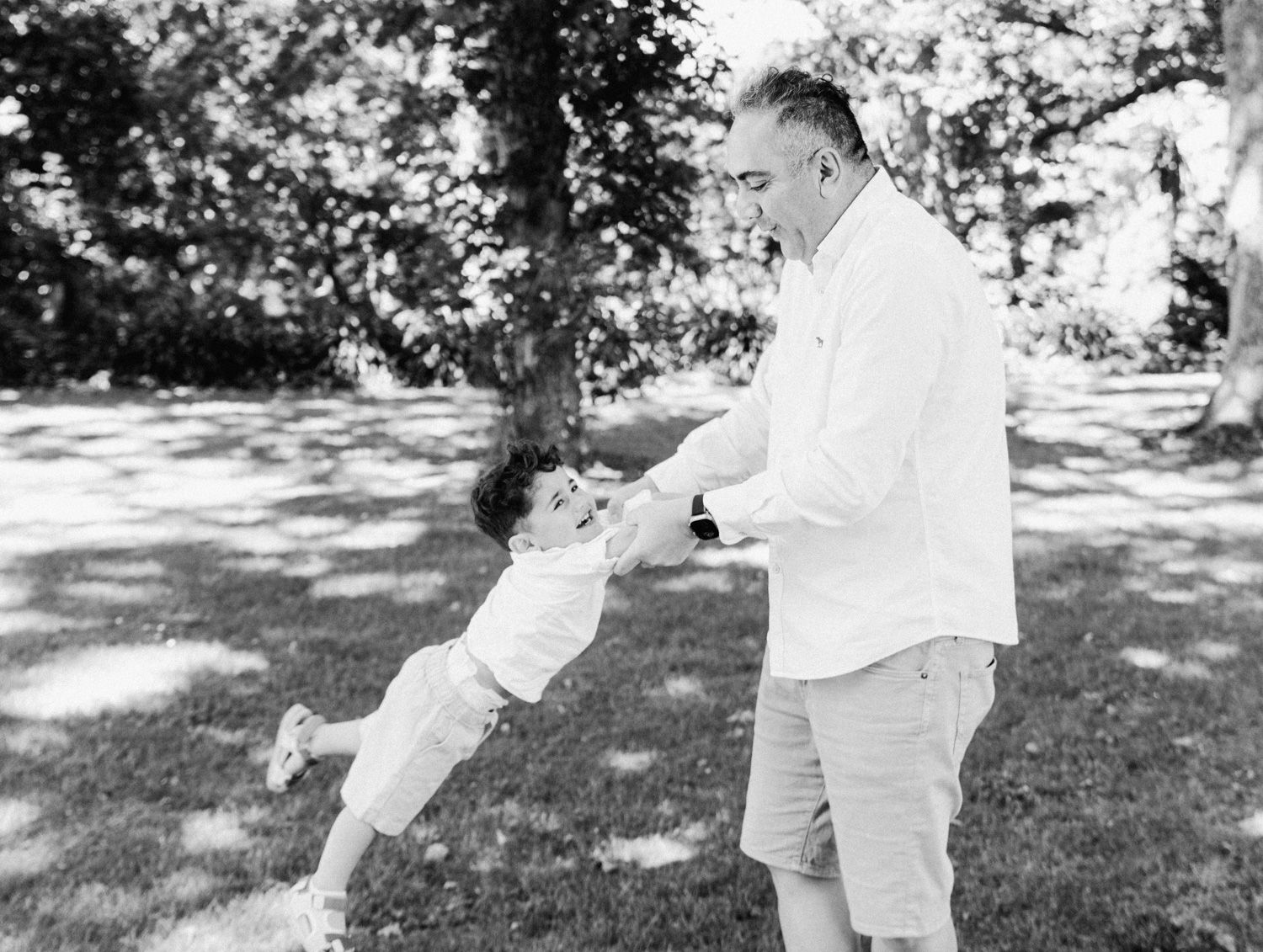 Black and white photo of a father joyfully swinging his young child in a sunny park surrounded by lush trees.