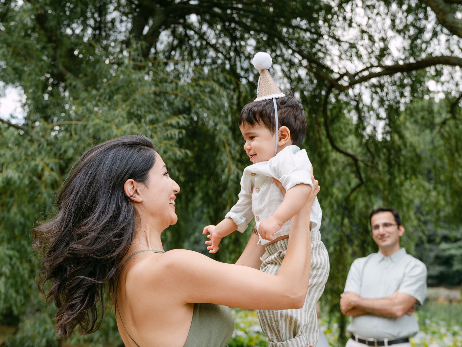 A mother joyfully lifts her toddler wearing a birthday hat while a man smiles in a lush green outdoor setting.