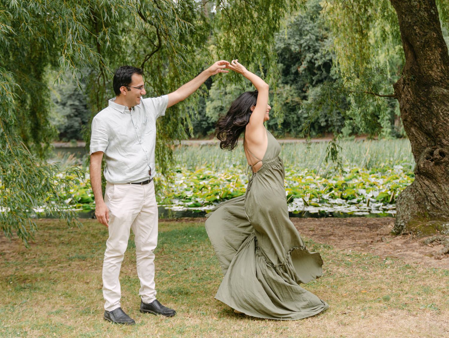 Couple dancing outdoors under a willow tree in a lush garden, woman in flowing olive dress being twirled by man in white.