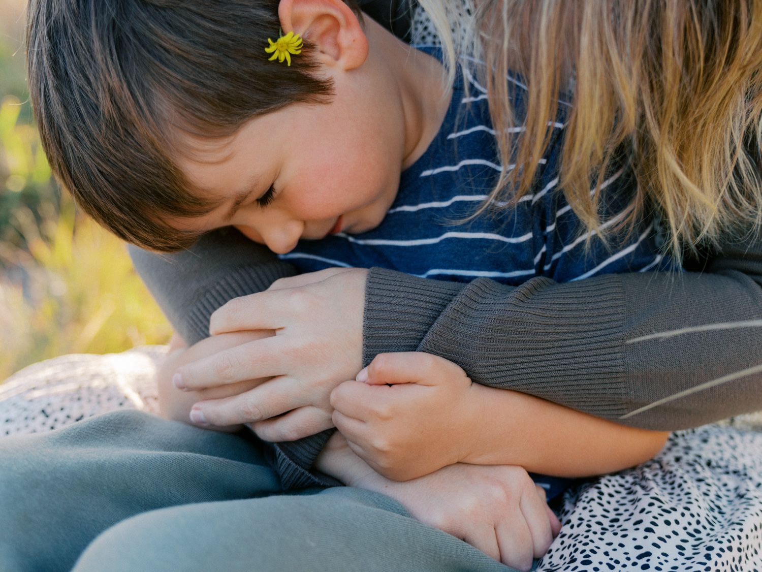 A young child with a flower behind their ear cuddles closely with an adult outdoors in a tender embrace.