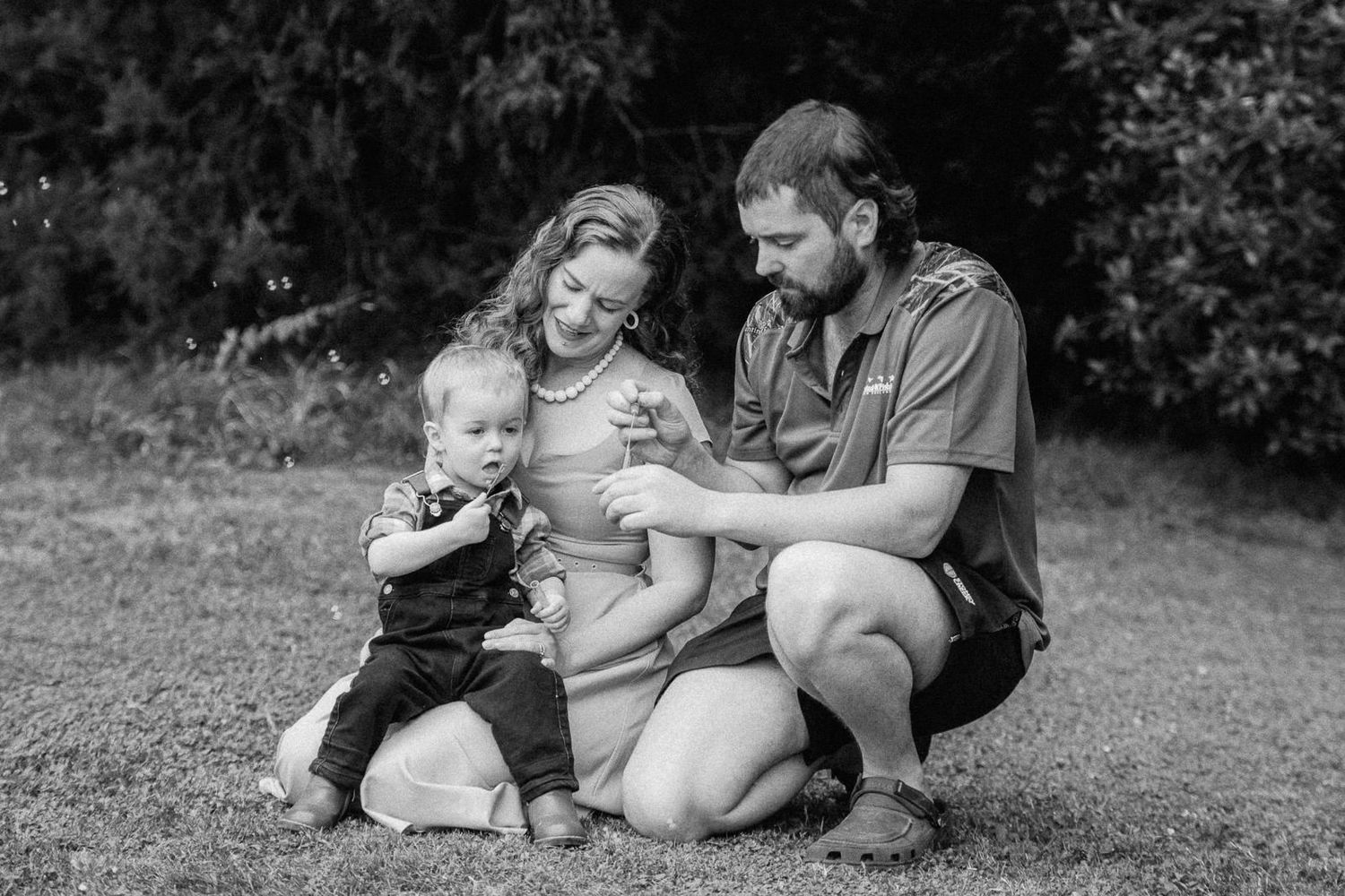 Black and white family photo of parents and toddler sitting on grass with green trees in background.