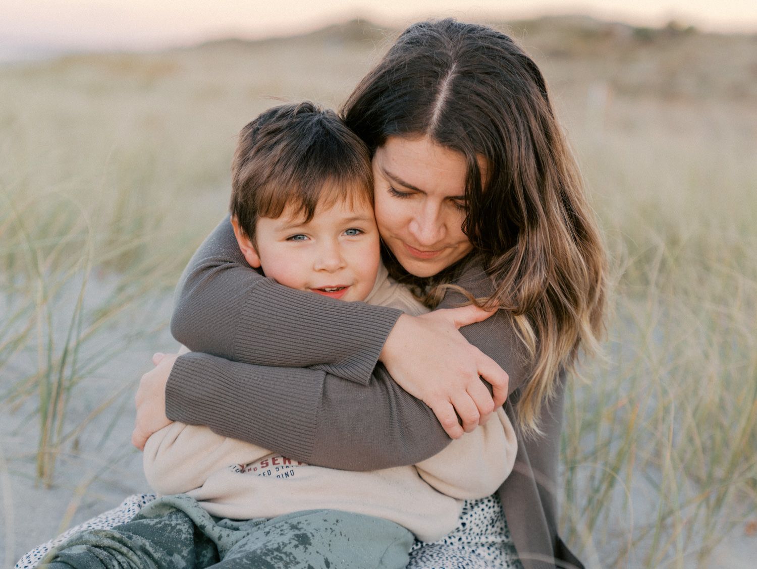 A mother tenderly hugs her young son on a sandy beach with dune grass in soft golden light.