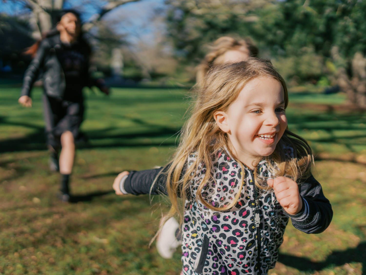 Happy young blonde girl runs with arms out in a sunny park, adult visible in background.