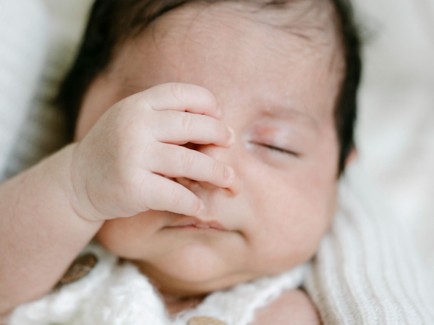 Close-up of newborn baby's face with tiny hand raised near eyes on soft white blanket.