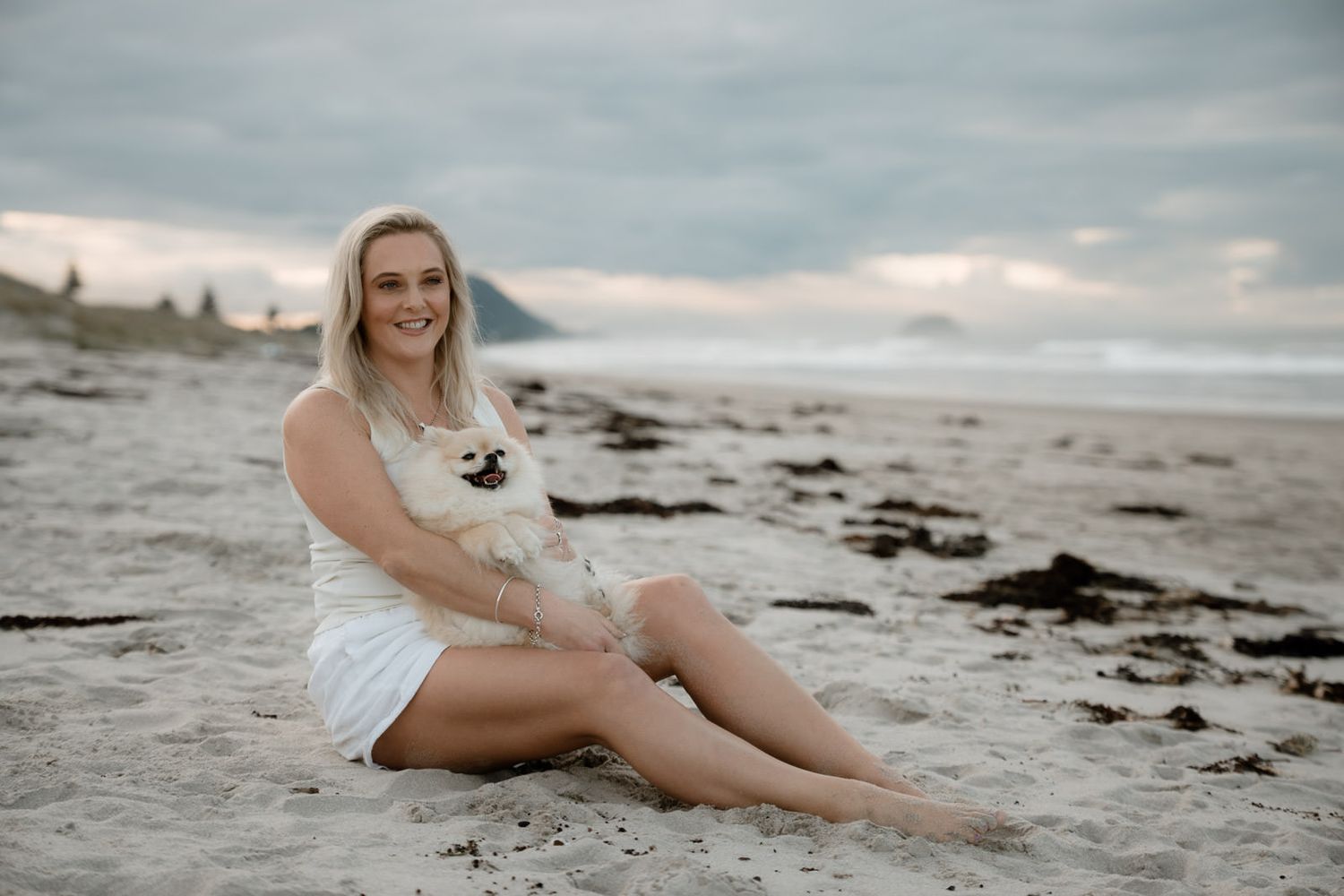 Smiling blonde woman in white dress sitting on beach holding small white dog with cloudy sky backdrop.