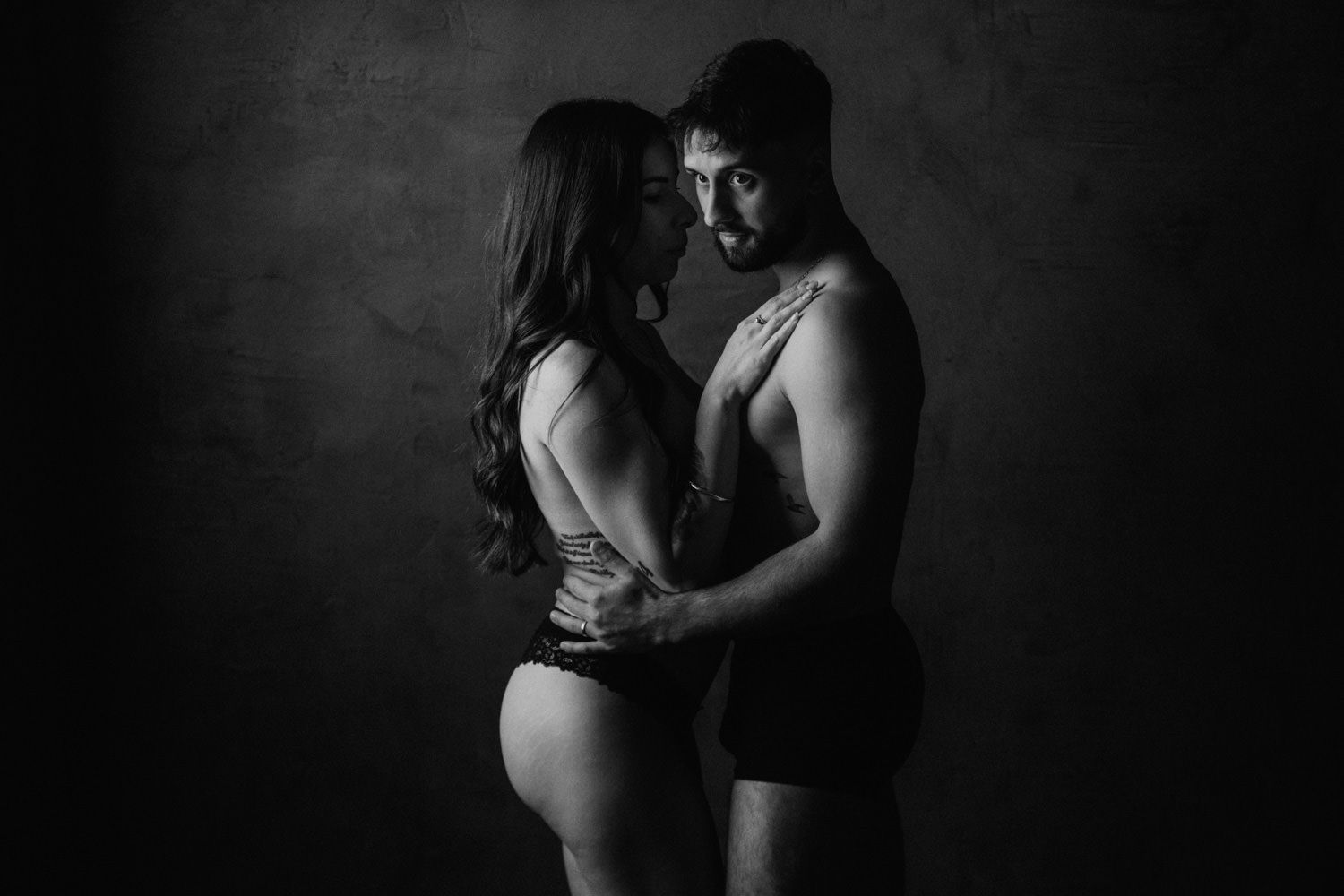 Black and white intimate couple portrait, man and woman in close embrace wearing minimal clothing against dark studio background.