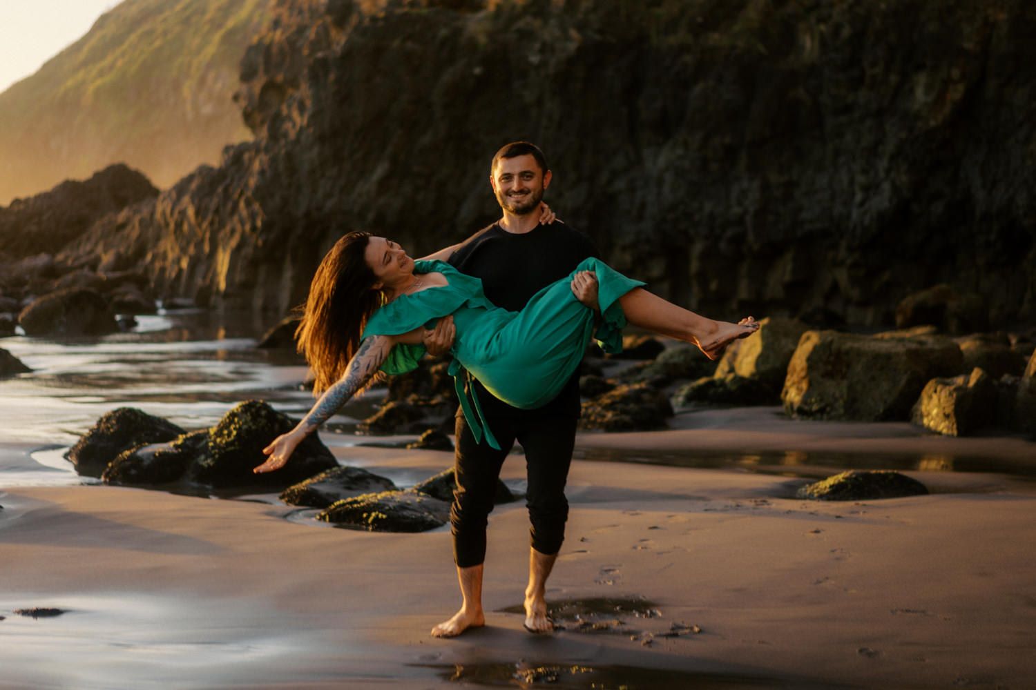 A man lifts a woman in a green dress on a rocky beach at sunset, posing playfully near mossy boulders.