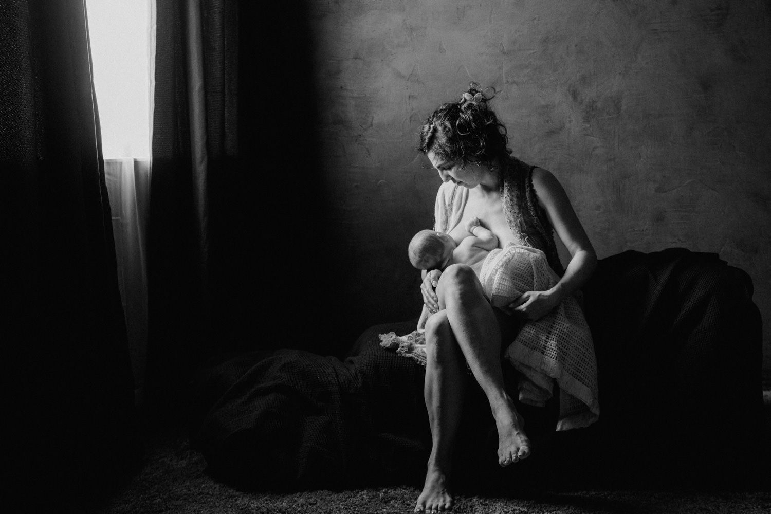 Black and white photo of a mother breastfeeding her newborn baby while sitting on a bed near a window.
