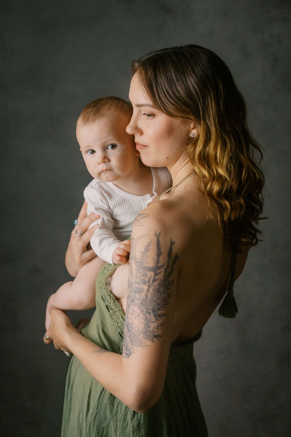 A young mother with a tattoo in a green dress tenderly holds her baby in a studio portrait.