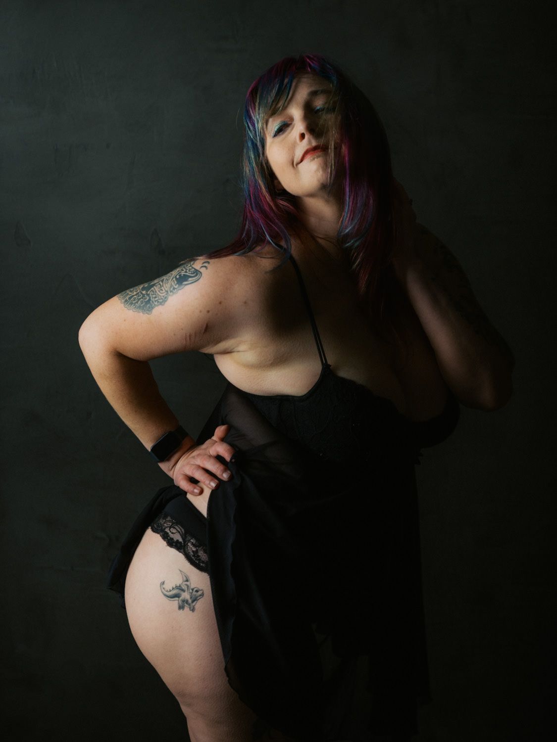 Woman with colorful hair in black slip dress poses confidently, showcasing tattoos on arm and thigh against dark backdrop.