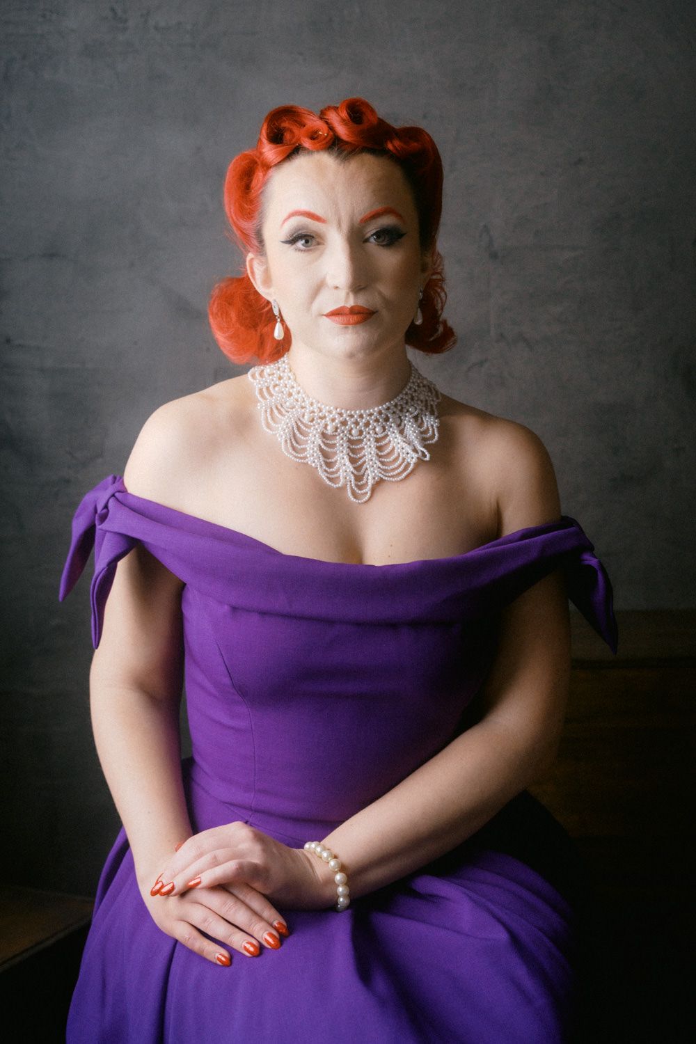 Woman in purple off-shoulder gown with red updo hair, pearl necklace, seated against gray background.