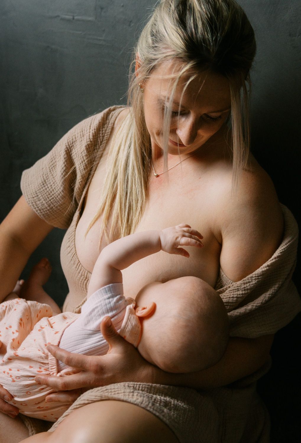 A blonde mother tenderly breastfeeding her newborn baby, wearing a beige textured top in a softly lit studio.